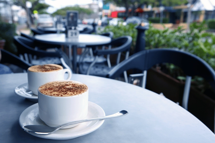 two hot coffees sitting on an outdoor table outside of a coffee shop